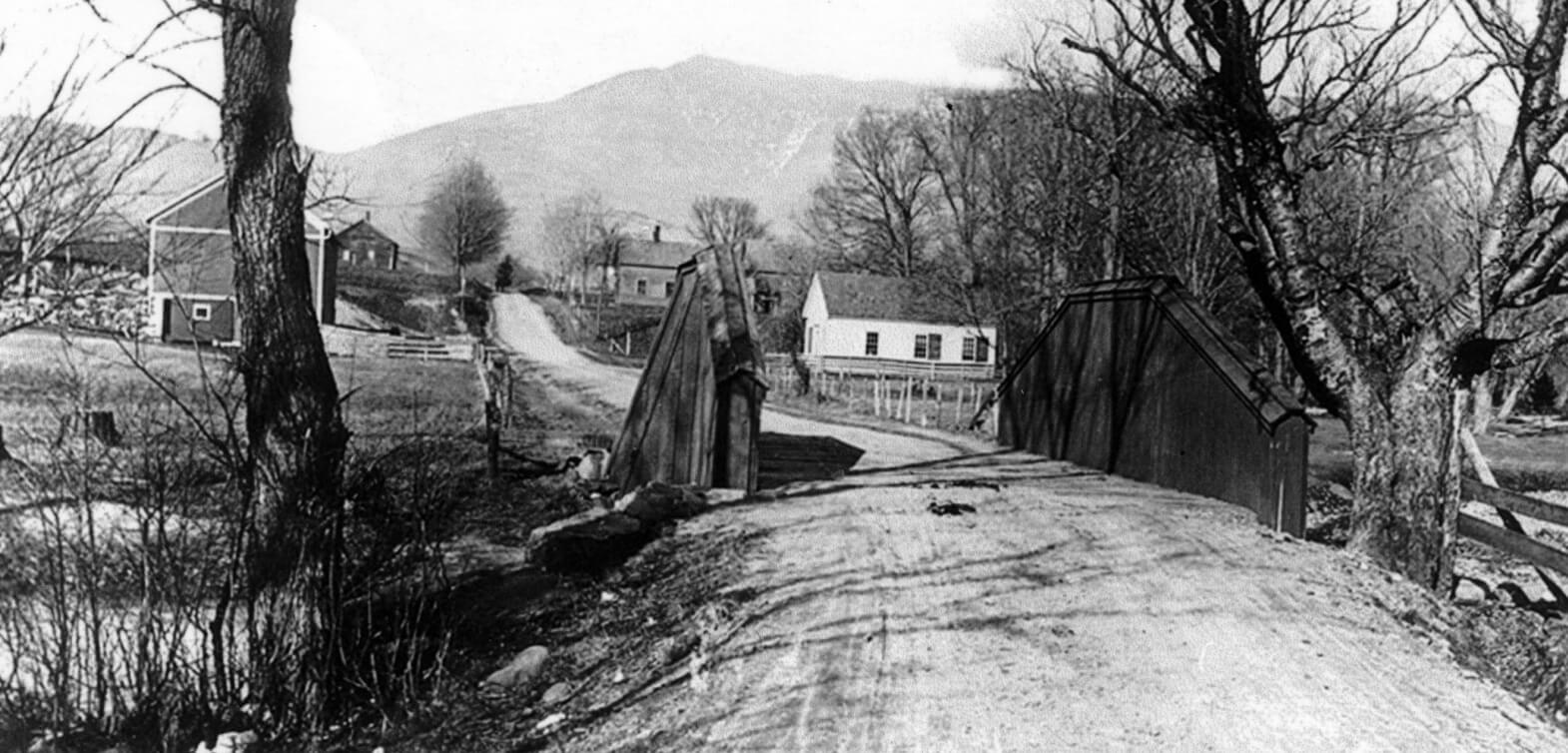 Old black and white photo of a Schoolhouse on a dirt road in the fall, maybe taken around the turn of the century.