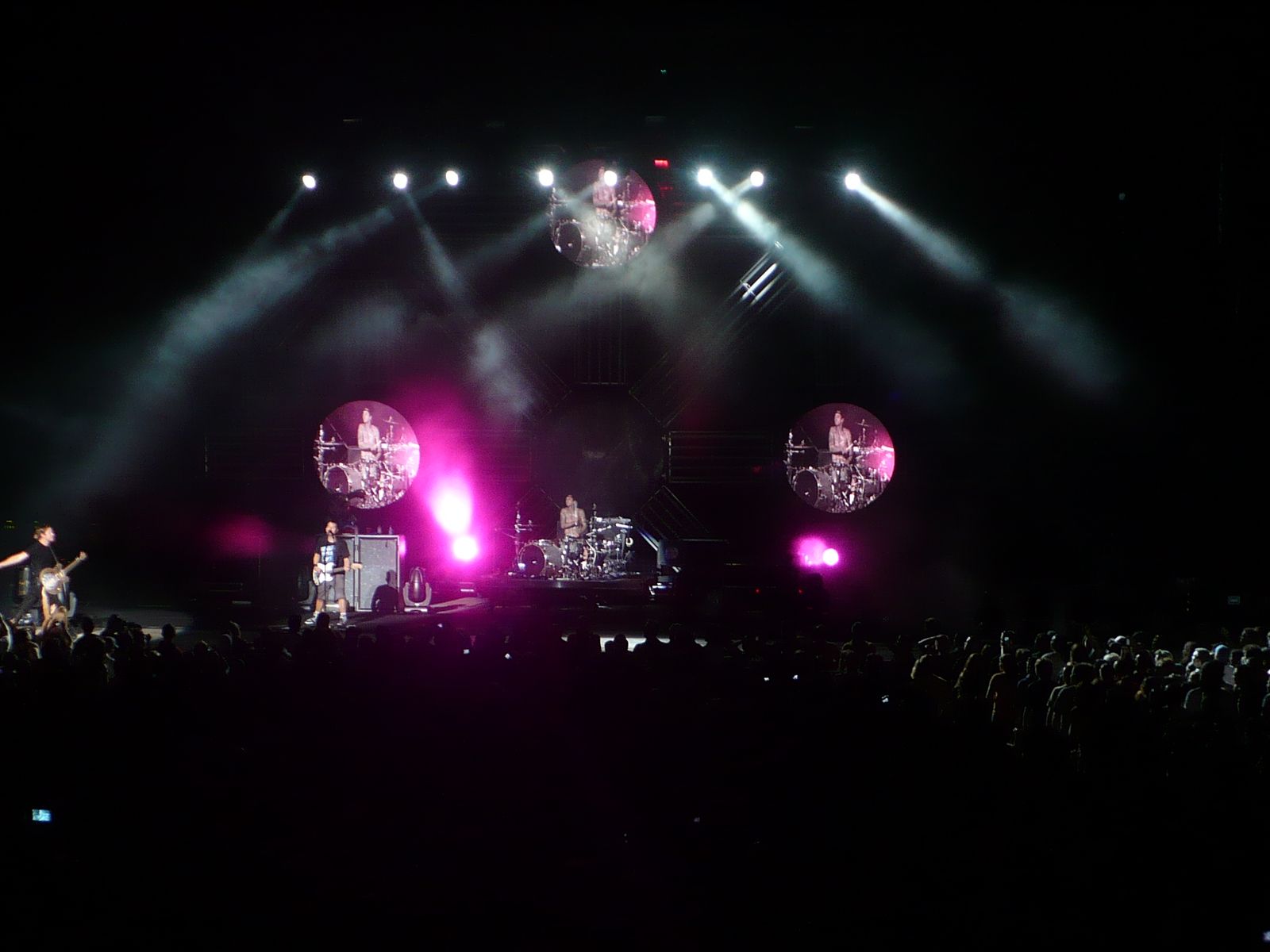 blink-182 performing on a massive amphitheater stage with three circular screens projecting Travis Barker drumming, pink stage lights and smoke