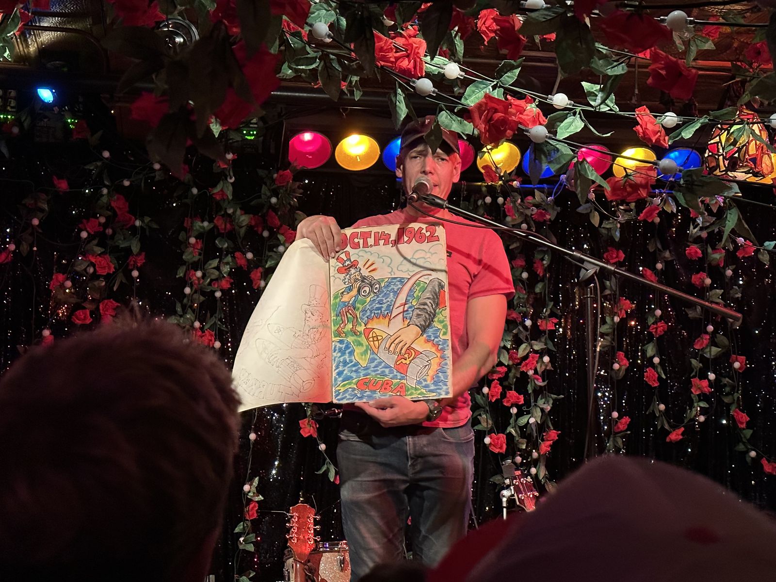 Jeffrey Lewis holding a comic panel reading 'Oct. 14, 1962' and 'Cuba' as he narrates the crisis through song