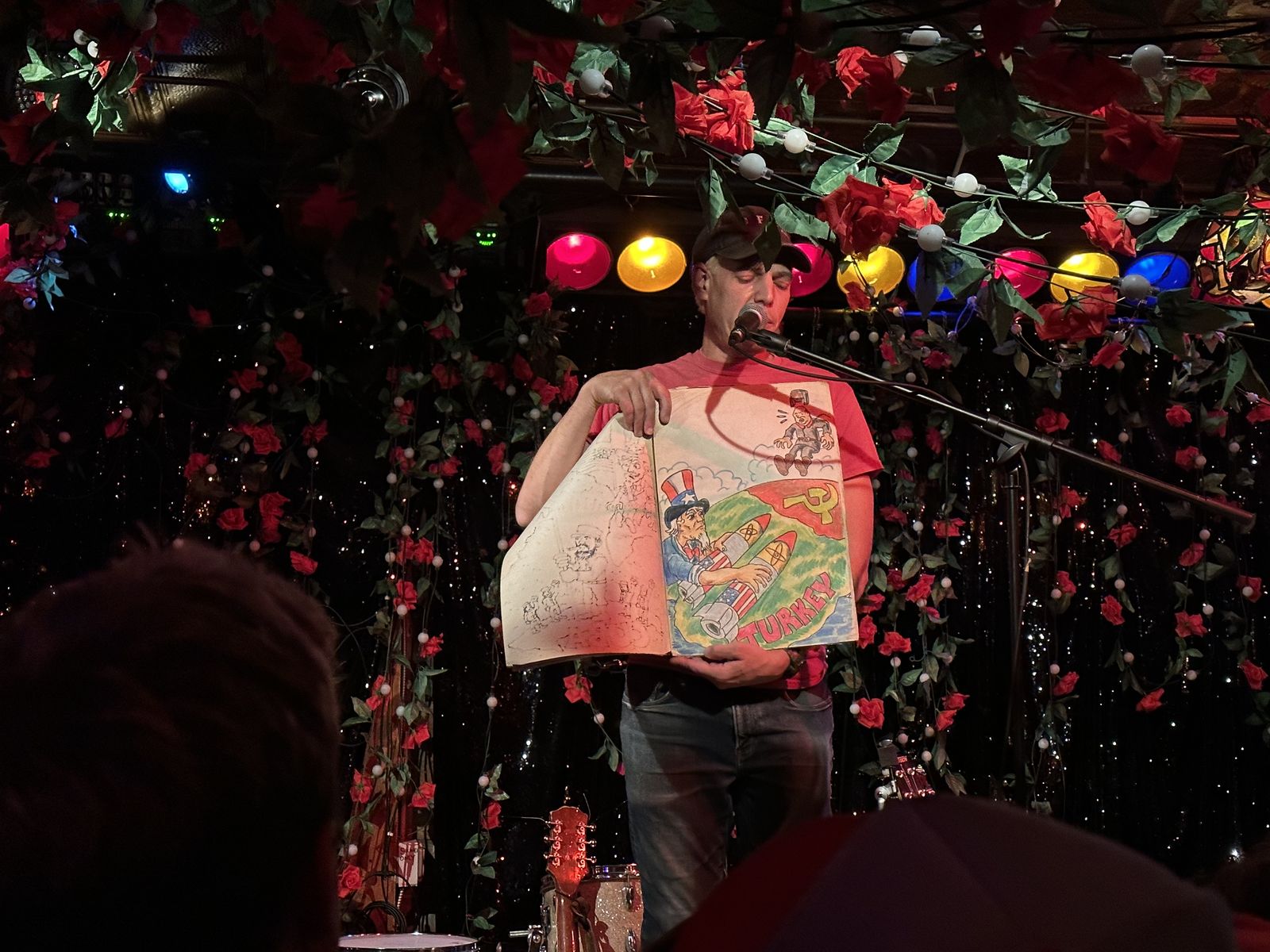 Jeffrey Lewis mid-performance holding a comic panel showing Uncle Sam and a Soviet symbol with 'Turkey' written on it, part of the Cuban Missile Crisis song