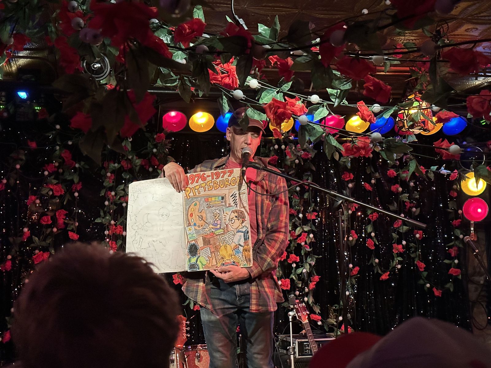 Jeffrey Lewis holding up a large hand-drawn comic book titled 1976-1977 Pittsburgh, performing one of his non-fiction comic songs