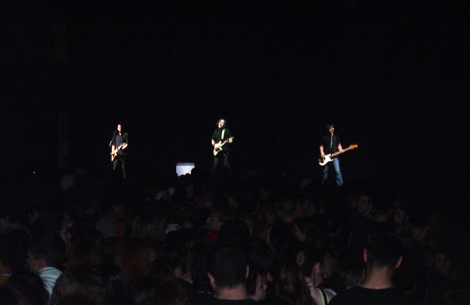 Jimmy Eat World performing, three guitarists spread across a dark stage with crowd in foreground