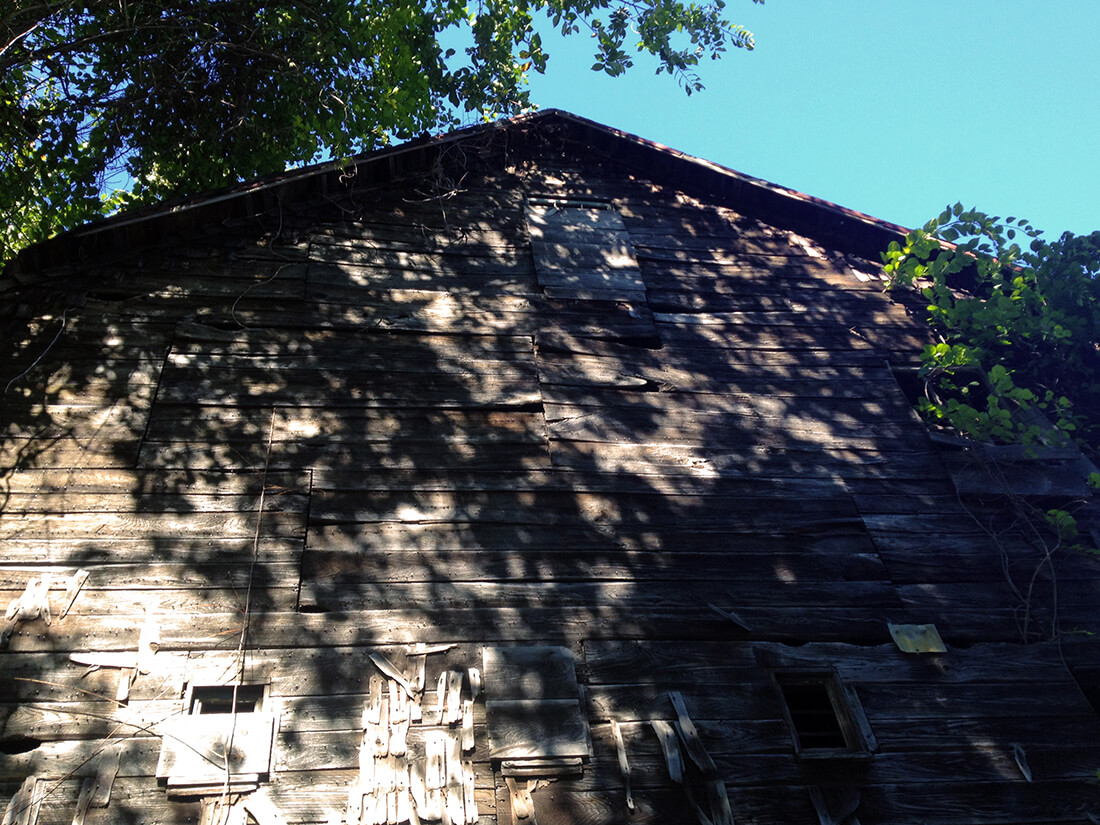 A brown and patched barn coveredwith shadows from unseen trees