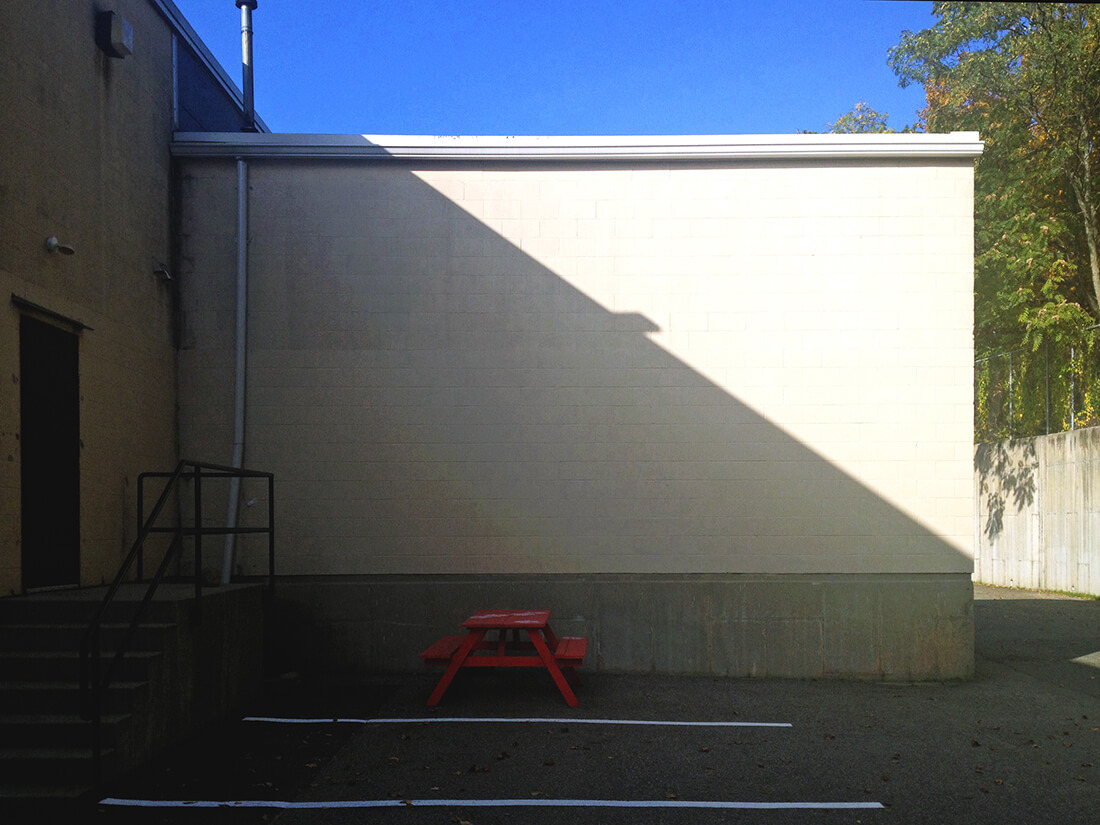 Harsh shadows in a nook behind a strip mall with a red picnic table in the center