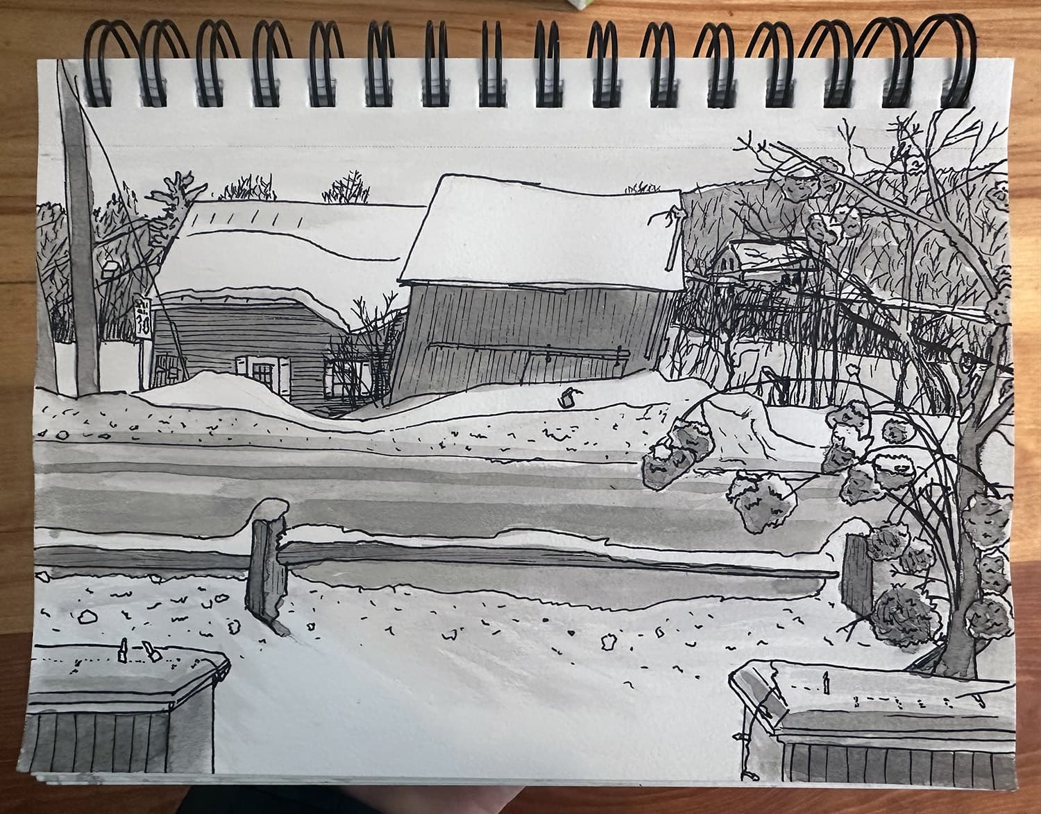 pen and inkwash sketch of the posts from a porch in the foreground, a snowy road, and two barns across it covered in snow. one barn is tilted but still standing, trees in the background.