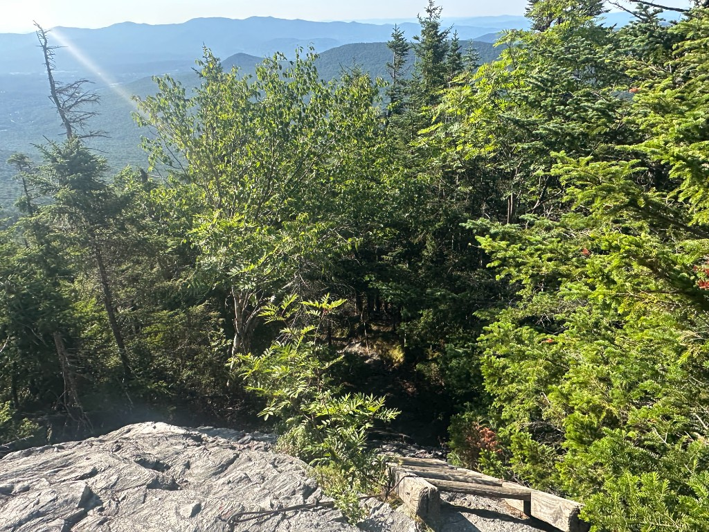 Looking straight down a steep granite ledge drop, spruce trees and hazy ridgeline below