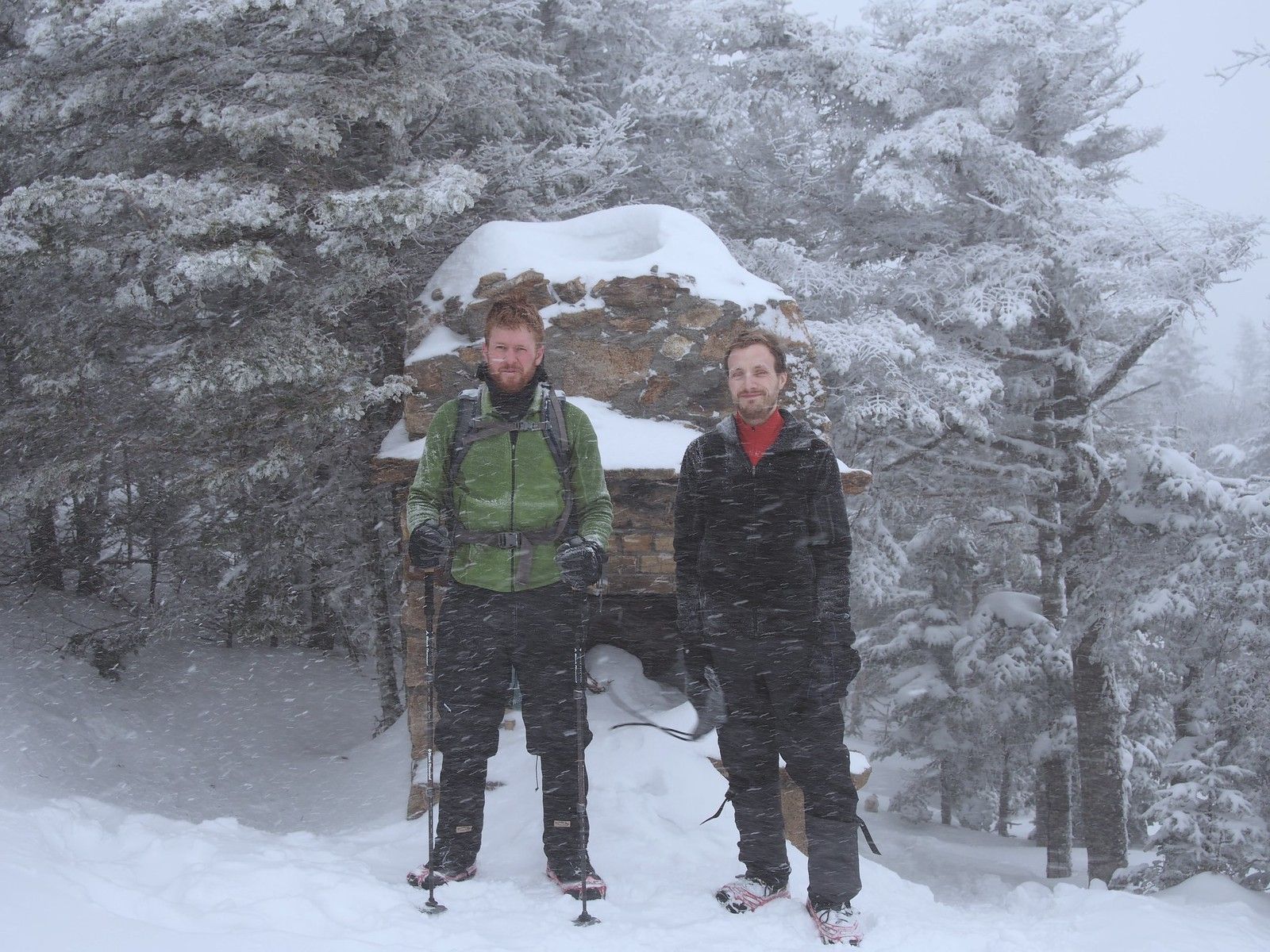 Two hikers squinting through the snow for a picture at the top of a snowy mountain